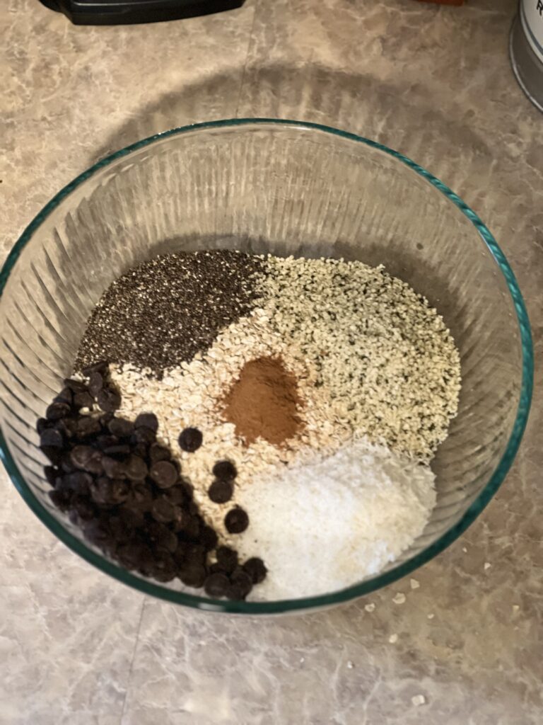 Mixing bowl sitting on a counter top containing oats, chia seeds, chocolate chips, shredded coconut, and ground cinnamon. 